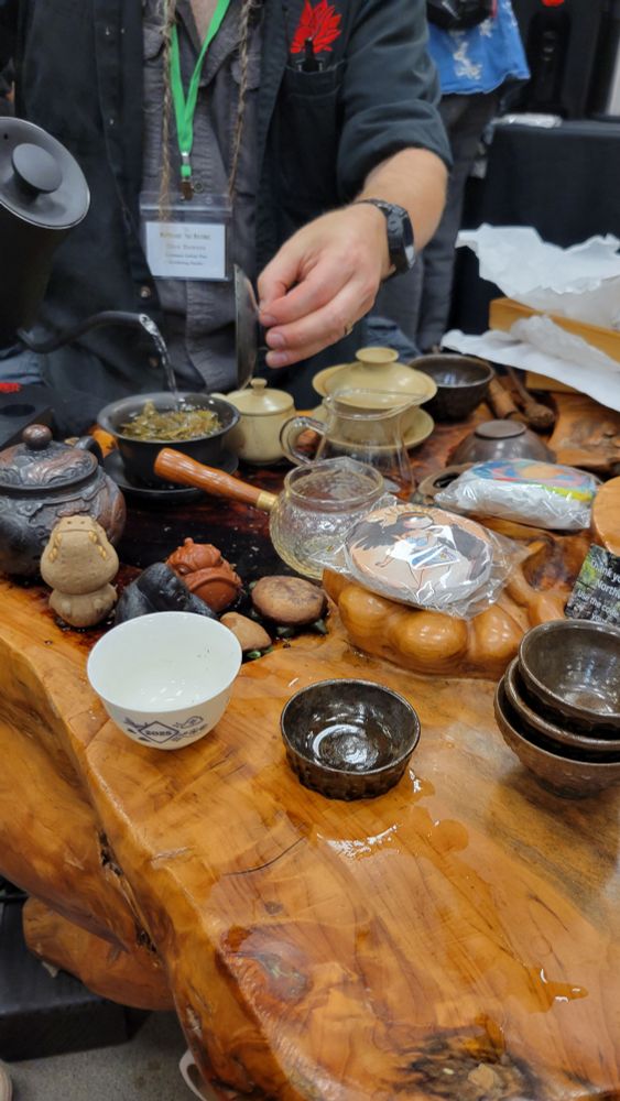 Picture of a tea table with several tea pots, gaiwans, cups, and tea pets; with trays of tea, and water being poured from a kettle into a gaiwan.