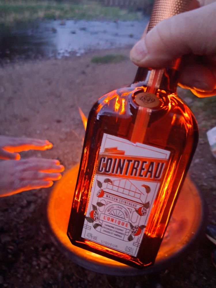 A glass bottle of the orange flavoured liqueur, Cointreau, held in  front of a campfire. Someone's hands are warming from the glow of the fire.