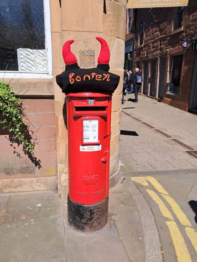 A pillar box style post box in the town of Kirriemuir, Scotland but with a knitted cover on top. The cover says Bonfest and has horns to celebrate the rock band ACDC