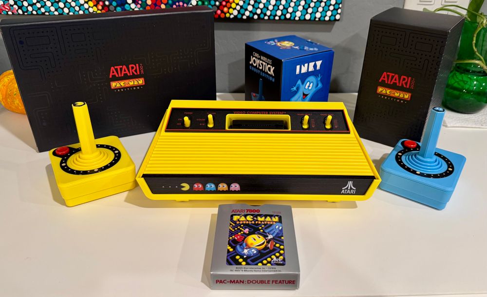 A bright yellow “Pac-Man edition” Atari console with a matching yellow joystick and a medium blue joystick. There’s a picture of Pac-Man, with red, blue, orange, and pink ghosts chasing him on the front of the console. They light up when it’s powered on. 