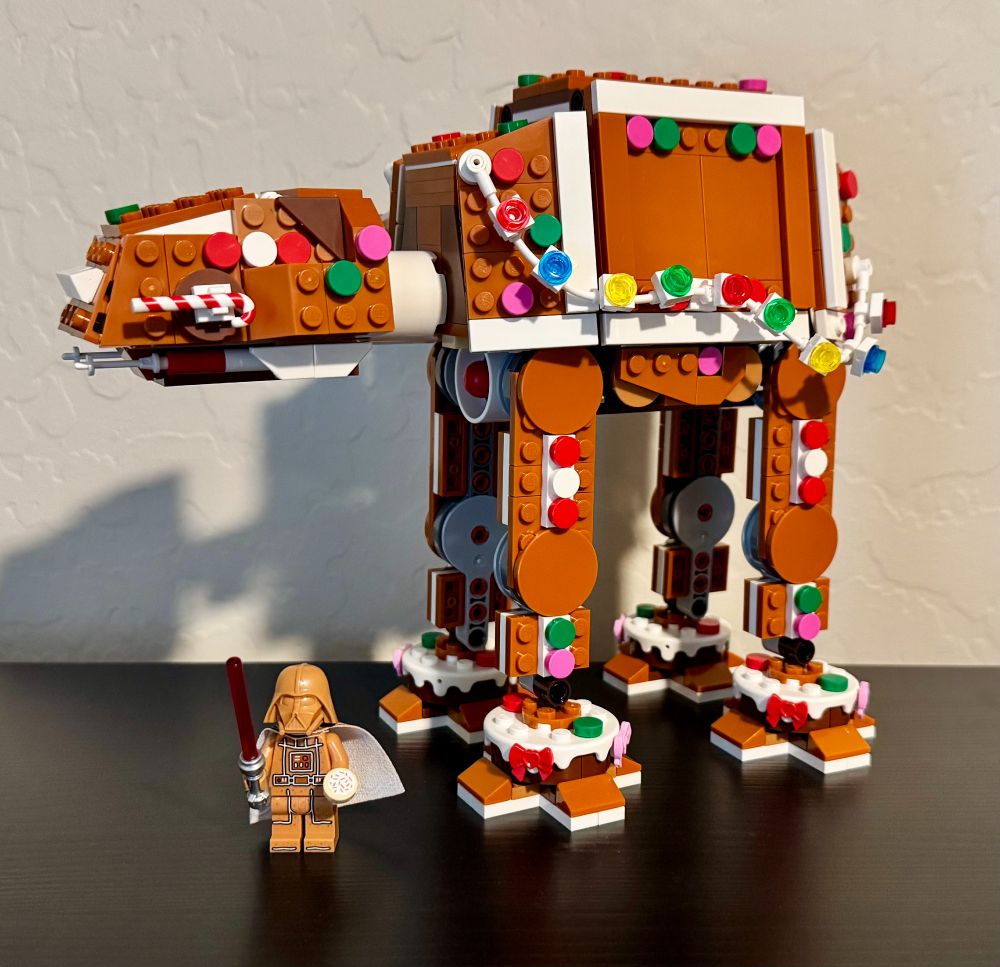 A LEGO model of a Star Wars AT-AT that looks like it's made out of gingerbread for the holidays. The model is decorated with a string of LEGO Christmas lights in red, blue, yellow, and green, candy canes, and other holiday items (the lights are not functioning, just decoration). There is a minifigure included of Darth Vader that is also a gingerbread version (light brown color with white frosting details).