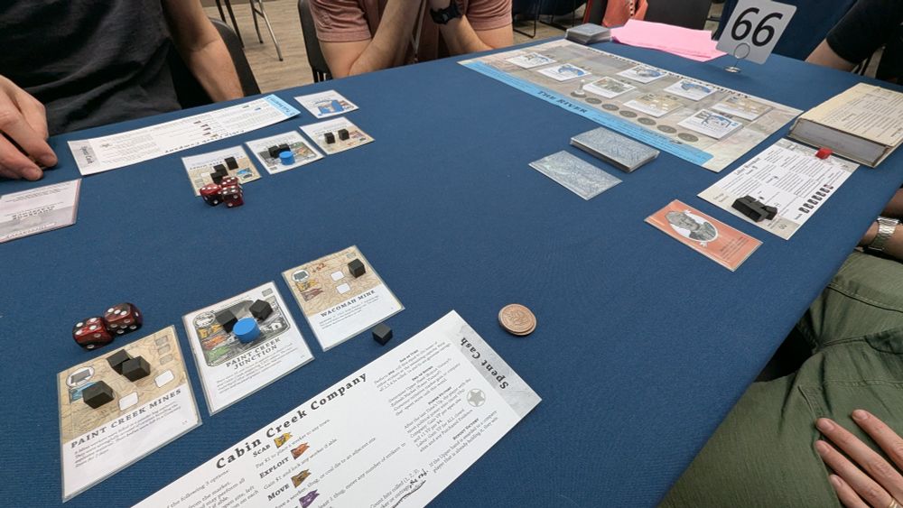 A table with a board game Hell Raisers in Kanawha County set up, with cards, dice, cubes, and boards seen on the tablet. A standee indicates that table number is 66.
