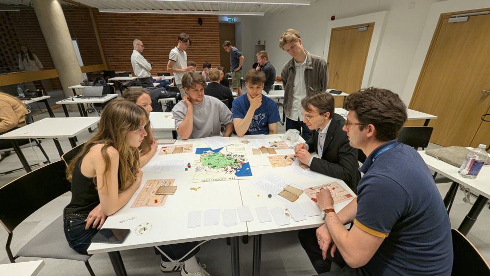 Six people sitting around a table, and playing a game. The map depicting the Grand Duchy of Lithuania is in the centre. Each player has additional cards and boards. There are units on the map.