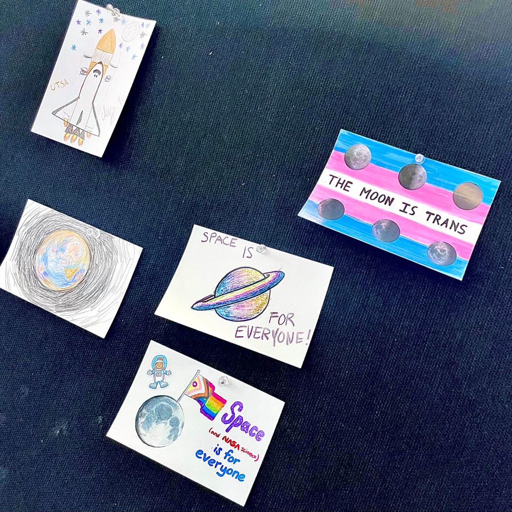 A close-up photograph of the “artistic creations board “at LPSC 2025. It is a black pushpin board, with no cards that participants have colored, written on, placed stickers on. there are five cards in this scene, and three of them are about LGBTQ people. The top one, and my favorite, is a trans pride flag in blue pink and white, with some moon stickers in a row on the top and bottom, and in the middle, it says “the moon is trans“. In the middle is a placard that says space is for everyone next to a rainbow colored ringed planet. Below that is a sketch of the moon with a giant inclusive pride flag sticking out of it, and the text “space ( and NASA science) is for everyone.”