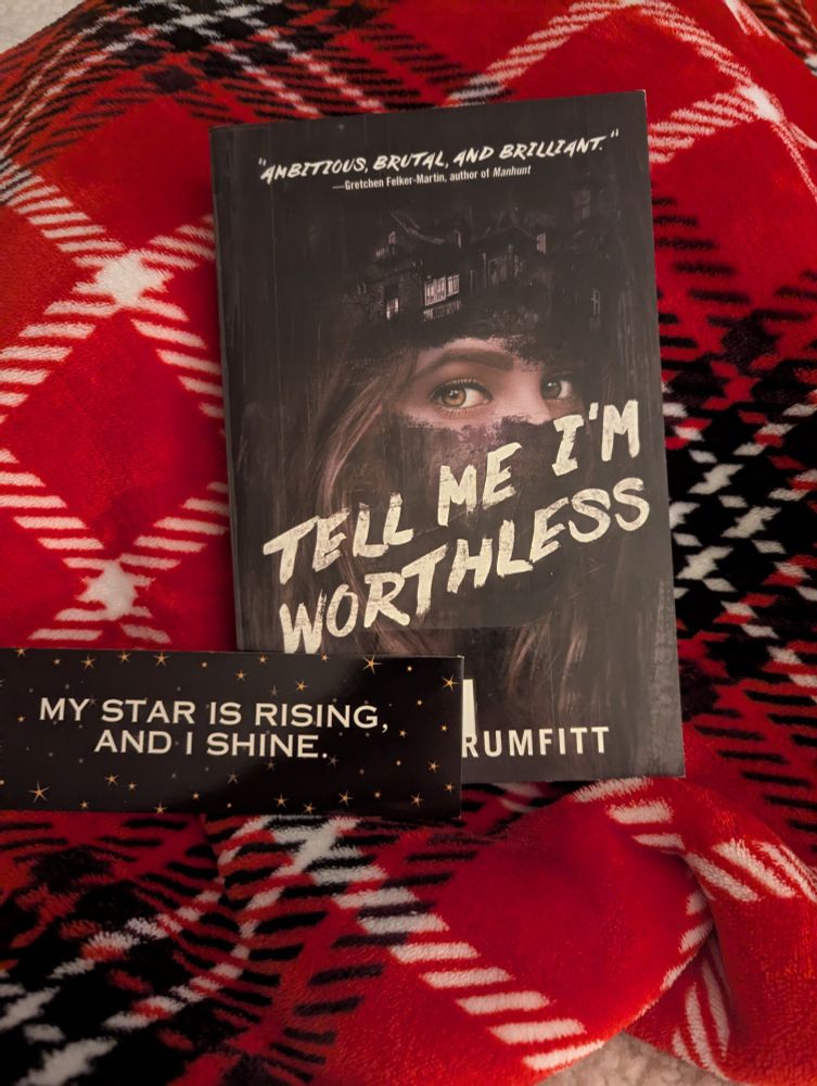 Cover of Tell Me I'm Worthless by Alison Rumfitt. Dark cover with a woman's partial face. House on her forehead. Book mark that says "my star is rising and I shine." Everything on top of a red, white, black plaid blanket. 