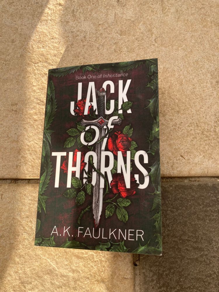 A photo of the front cover of A.K Faulkner’s Jack of Thorns novel. The cover is moody and atmospheric with the stark white title standing out against spiky foliage. In the center of the page is a sword, entangled in roses and thorns. 