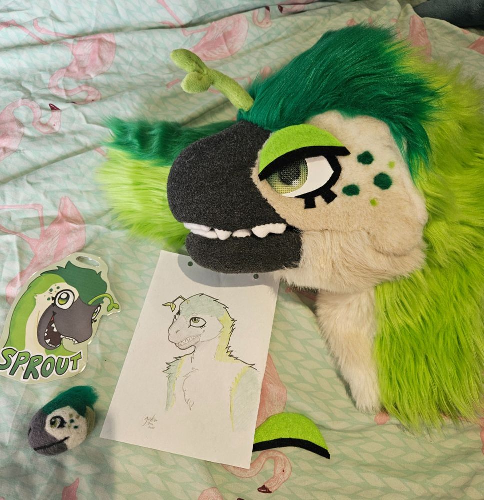 A photo of a dinosaur fursuit head with angry eyelids added. Added next to it is physical art of the character; a laminated badge, a pencil drawing, and a needle-felted head