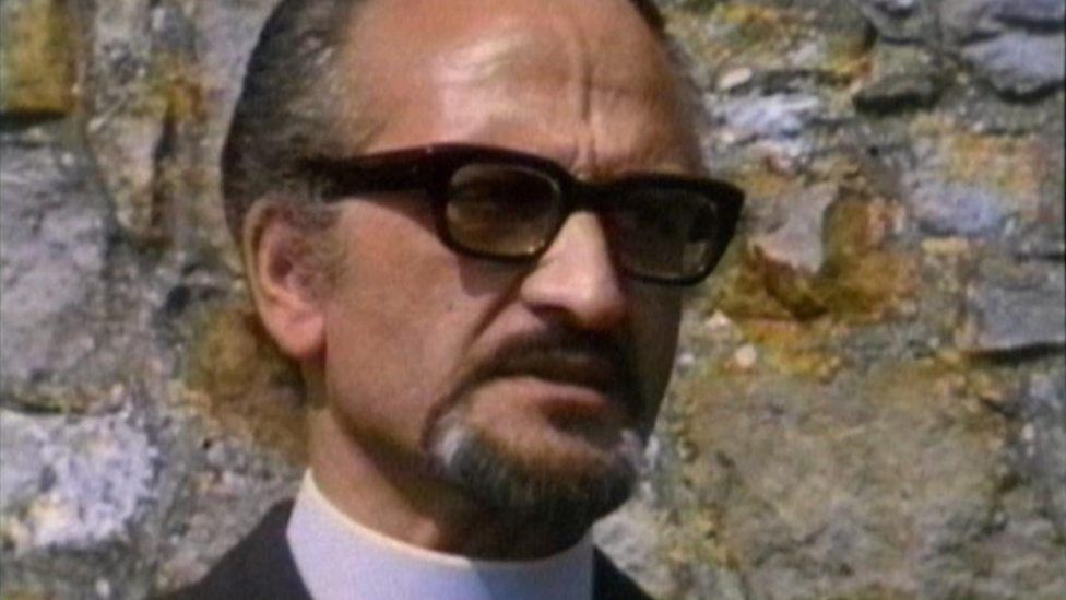 A goateed figure in a priest’s collar wearing stylish eyeglasses.