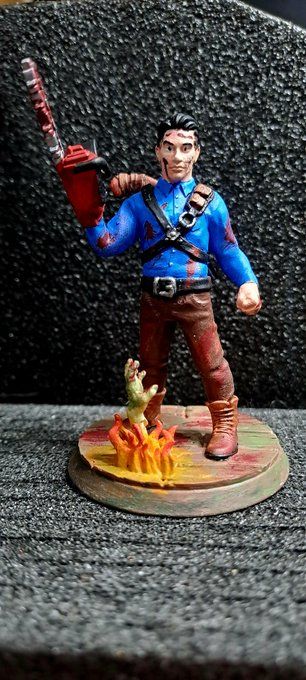 Miniature 3d model of a man in a blue shirt, brown trousers. With a chainsaw as his right hand and shotgun on his back. While a fiery zombie hand reaches out of the ground. 