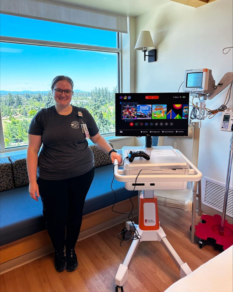 A Child Life Specialist stands beside a GO Kart gaming kiosk in a room with a large window view of trees and hills. The cart holds a monitor displaying a video game menu and a game controller. Medical equipment is visible on the wall. The person is smiling, wearing a dark t-shirt, and has a badge.