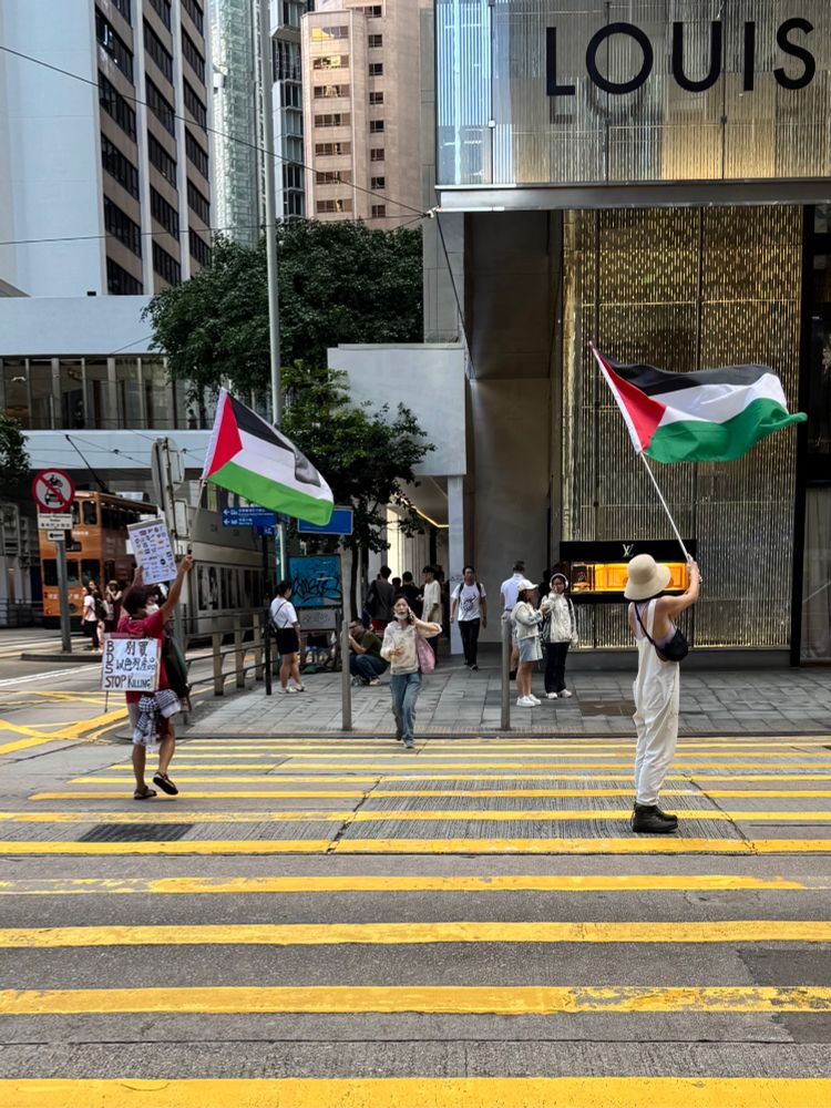Two ladies waving Palestinian flags on a crosswalk on Pedder Street, Hong Kong, with one carrying a placard urging a boycott of Israeli products