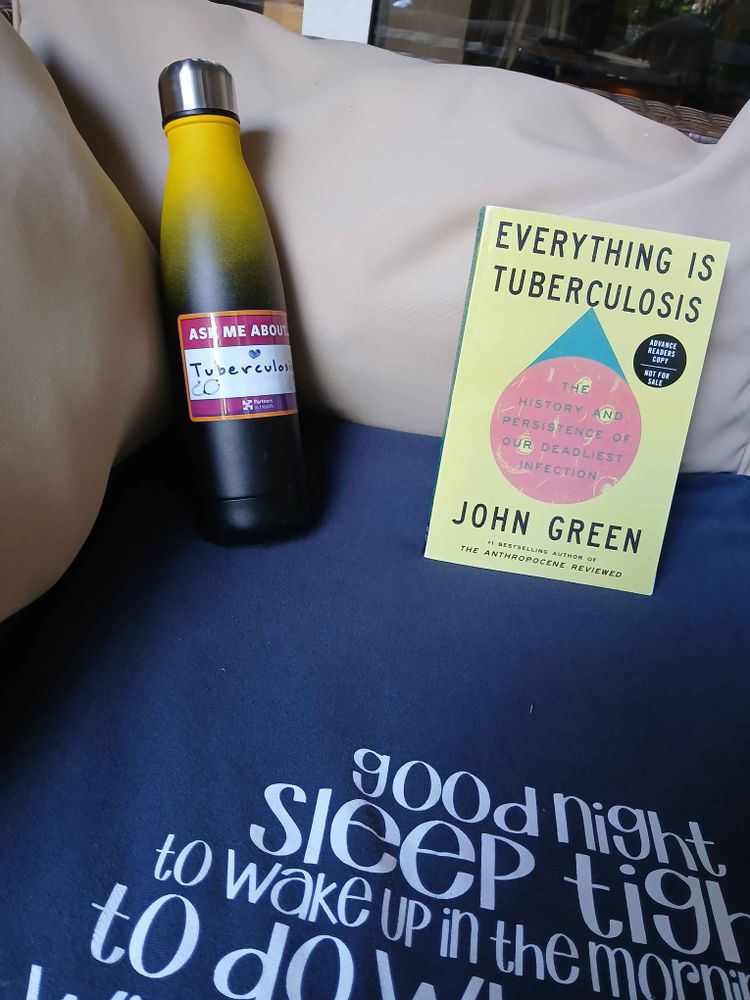 An advanced reader's copy of "Everything Is Tuberculosis" sitting on a blanket next to a water bottle with a sticker that reads "ask me about tuberculosis"