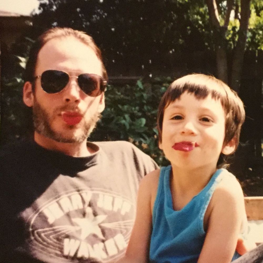 An early 90s photo of a father with his young kid on his lap in a backyard.  They both hold rose petals in their mouths so they look like red lips. The father wears a black tshirt and aviator shades with a short beard and slicked back hair. The kid is in a bright blue tank top with choppy hair and a barely contained smile that makes it difficult to hold the rose petals.
