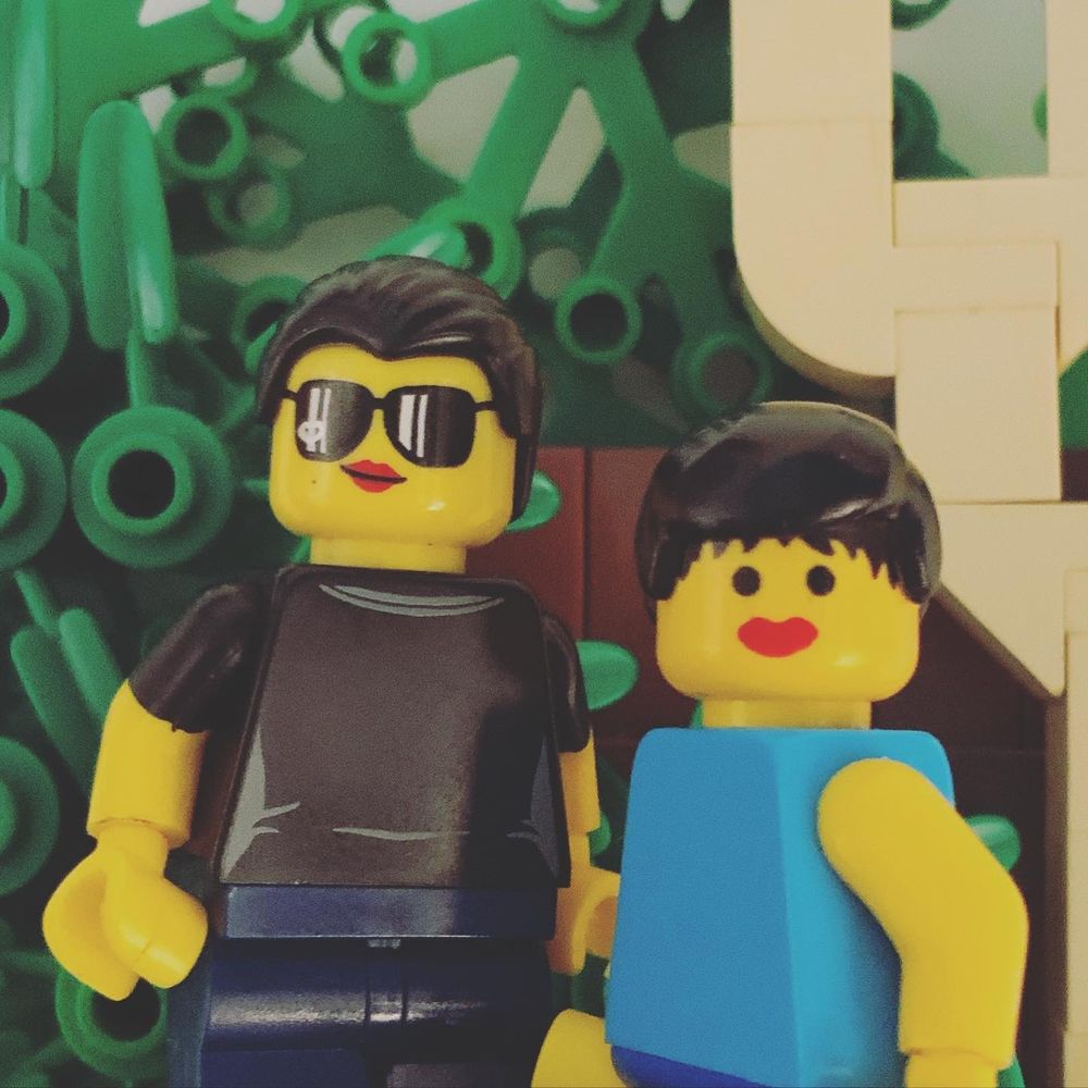 A recreation of the childhood photo in LEGO. Two minifigures stand in front of a wall of foliage pieces. They both use female-coded heads with red lipstick printed on. The only missing detail is the beard on the father’s face print.