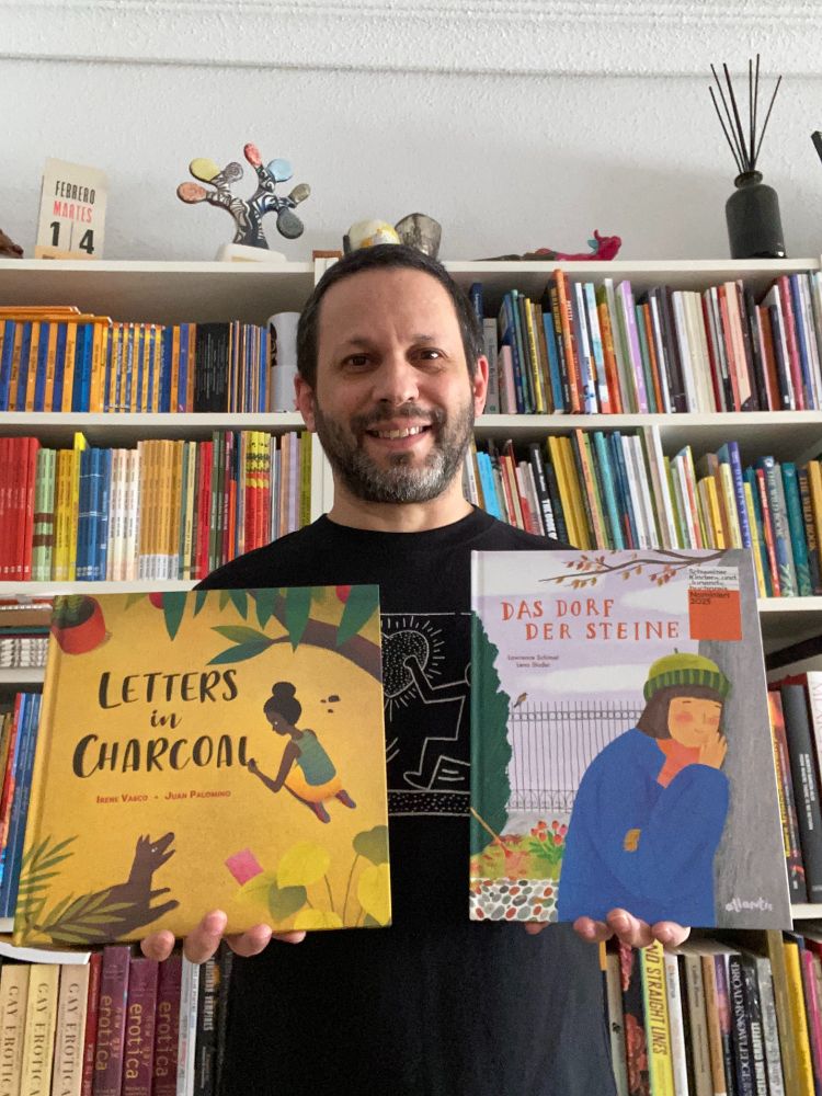 Selfie wearing a Keith Haring t-shirt standing in front of bookshelves of my own books or books I translated, holding 

in my left hand my picture book DAS DORF DER STEINE illustrated by Lena Studer, translated from Spanish to German by​ Eva Roth (Atlantis Verlag) which is a finalist for the Schweizer Kinder- und Jugendbuchpreis in Switzerland

&

in my right hand my translation into English of LETTERS IN CHARCOAL, written by Irene Vasco, illustrated by Juan Palomino (Lantana Publishing) which is shortlisted for the Carnegie Medal in Illustration in the UK.
