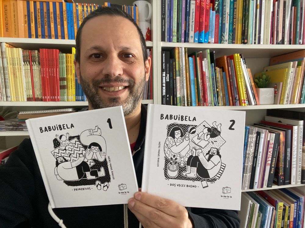 A selfie in front of a bookshelf, hold in one hand volumes 1 & 2 of Babuibela by Reesham Shahab Tirtho, square hardcover books with white covers each with a black and white pen and ink family scene (father, mother, little girl).