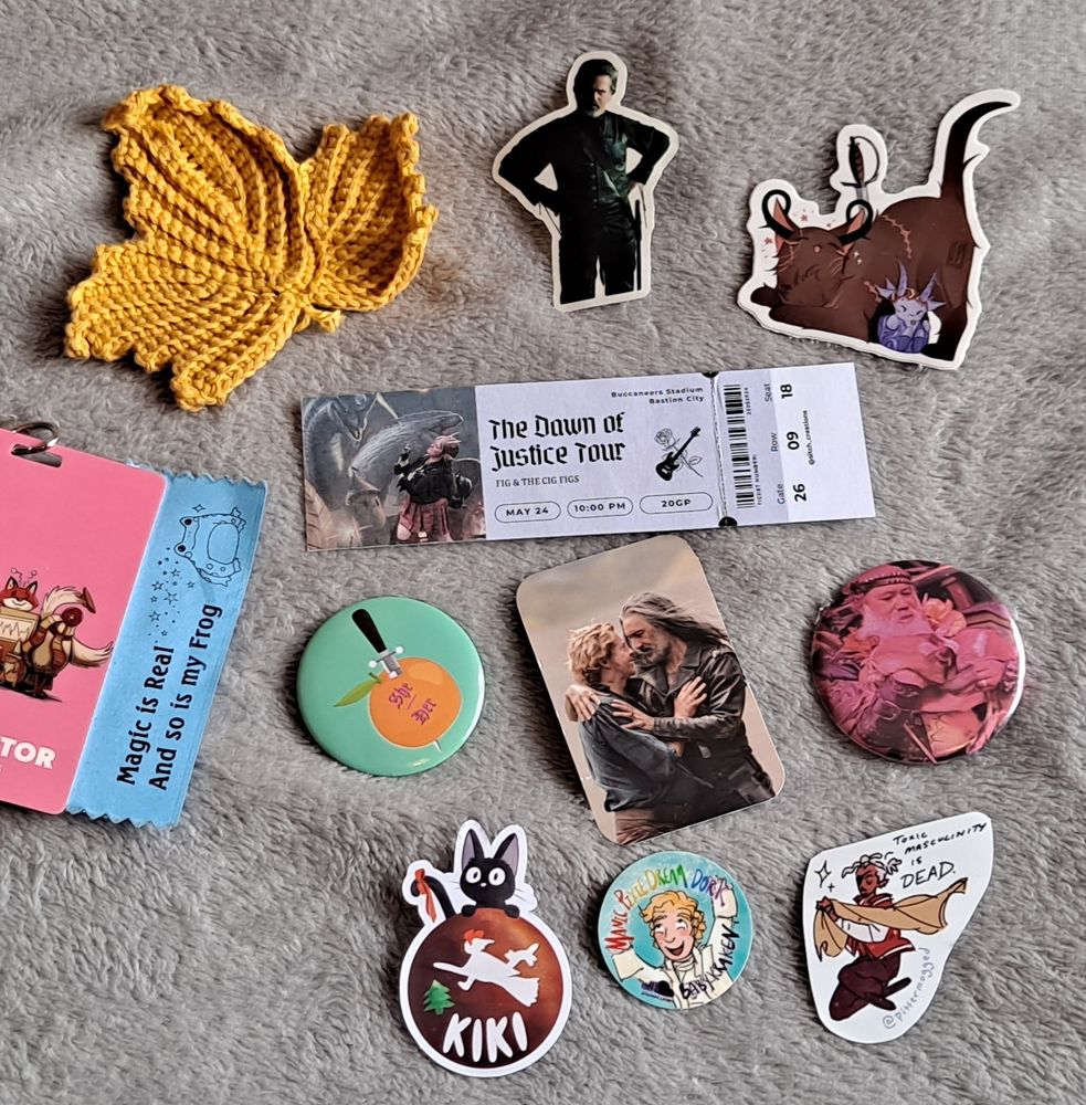 small items laid out on a blanket,  a crochet leaf, some stickers, a mirror, a button a fake band ticket and a photocard.