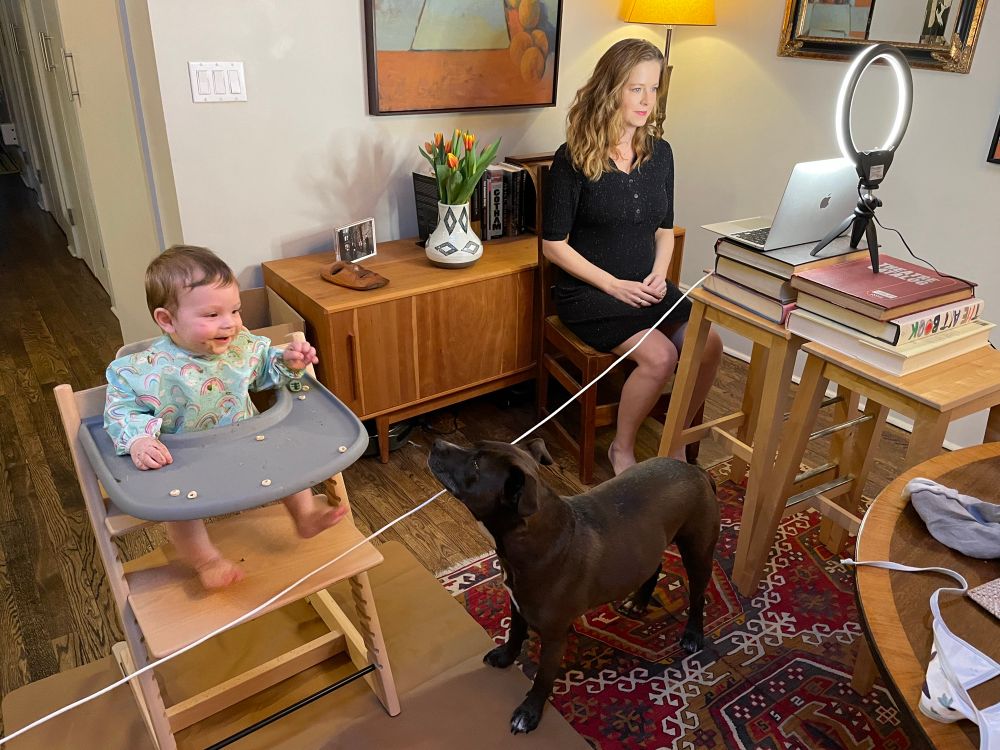 A woman about to go live on TV via zoom, while a baby eats cheerios in a high chair and a dog begs for crumbs. 