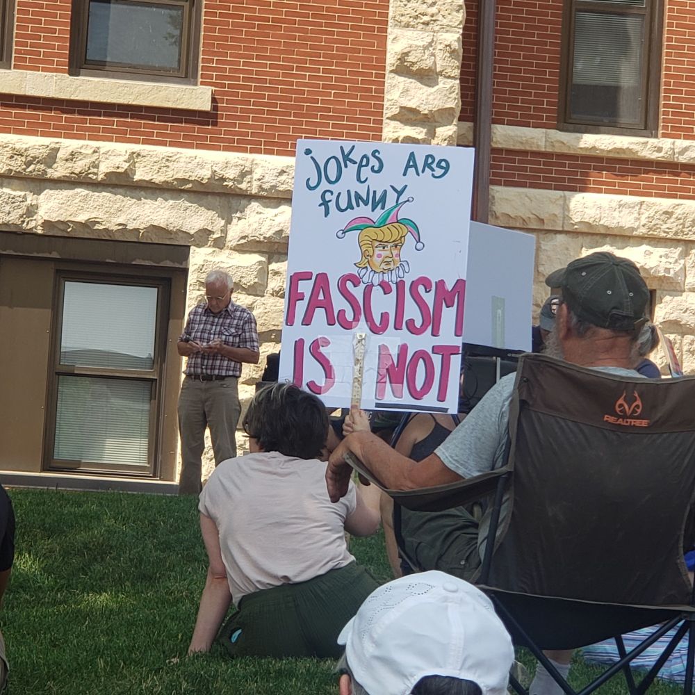 Foreground are people sitting on grass, one holds a political protest sign, in background is a large brick building 