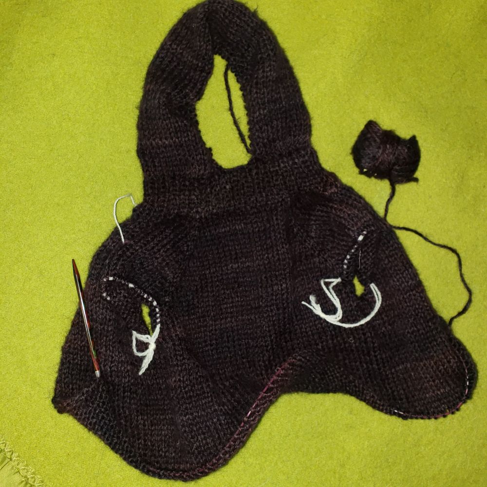 A partially knitted sweater lies flat on a bright green background. The yarn is shades of purple. Knitting needles can be seen on the bottom edge