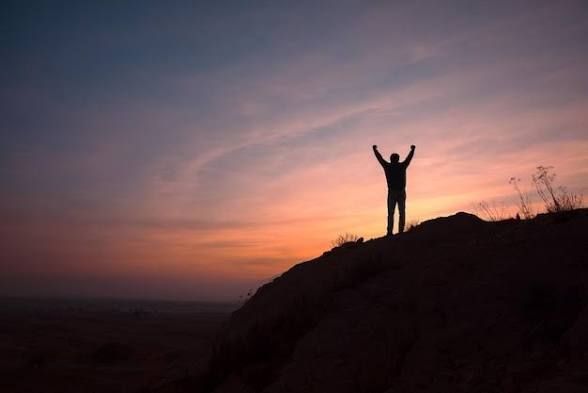 person standing on a rocky prominence, both hands in the air in a celebratory manner, sunset in the background
