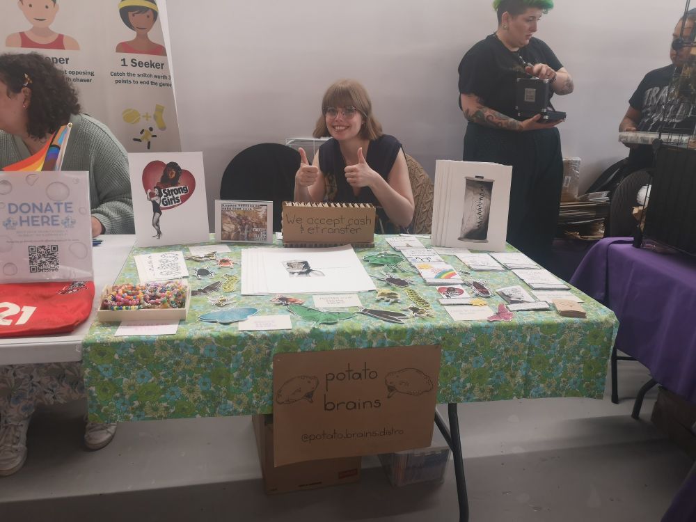 A light skinned blonde woman gives this up from behind a table with a green floral table cloth. The table is covered with zines and prints. There are 3 people in the background.