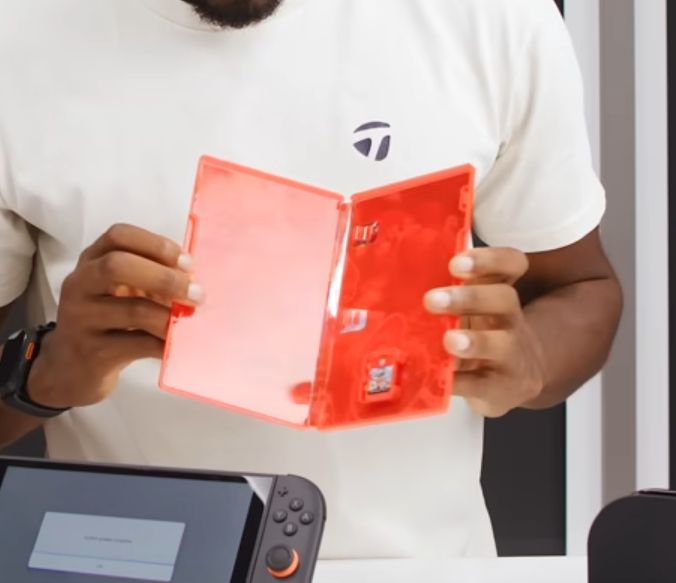 MKBHD in a white shirt holding an open red Nintendo Switch 2 game case, demonstrating the size contrast between the tiny game cartridge and the oversized packaging.