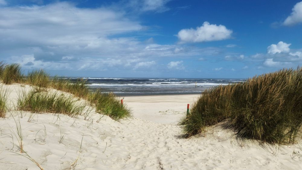 Ein Weg aus weißem Sand führt durch Sanddünen mit Dünengras hindurch auf einen Strand. Das Meer ist leicht wellig. Der Himmel ist blau, aber teils bewölkt. 