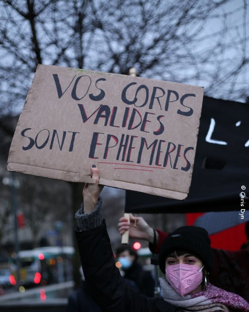 Manif à Paris, en hiver. Photo d'une jeune femme en masque FFP2. On ne voit que sa tâte et son bras. Le bras levé, elle tient une pancarte en carton sur laquelle est écrit : vos corps valides sont éphémères. 
