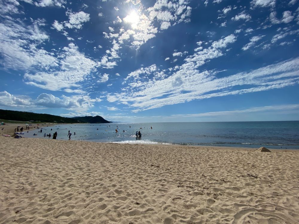 A photo of the Japan Sea coast, the top half showing deep blue sky, scattered puffy clouds of various shapes, and a bright sun; the bottom half shows the sea, beachgoers at a distance, and the curve of the sandy shore.