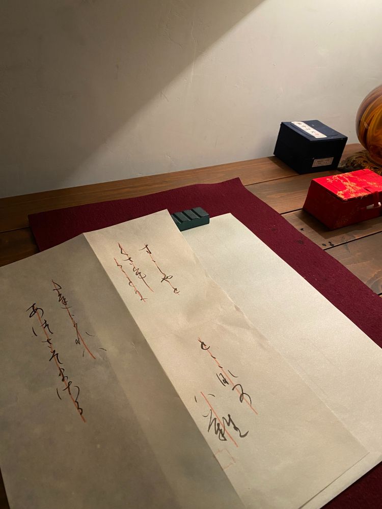 A photo of a vertical paper creased vertically in half, with a Japanese text written on it, intended to be copied as a calligraphy exercise. Transliterated into Roman orthography, the text reads "Suzushiya! to kusamura goto ni tachiyoreba / atsusa zo masaru, tokonatsu no hana" 