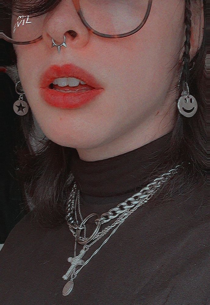 A close up picture of the bottom half of Moth’s face and their neck. They have several silver necklaces stacked around their neck, as well as two silver charms hanging from mini plaits at the front of their hair; one is a circle with a star cut out, and the other is a smiley face