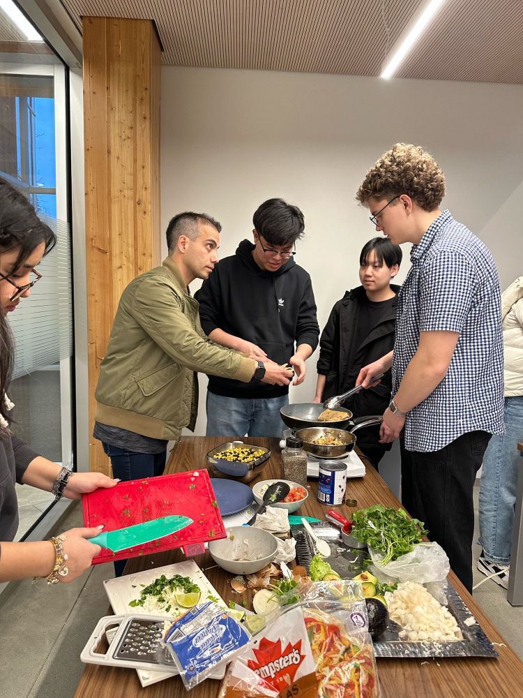 Students gathered around a table at a cooking event, preparing a meal together. Some are chopping ingredients while others cook on portable burners, surrounded by fresh produce and kitchen tools. The setting is a bright, modern indoor space with a warm, collaborative atmosphere.