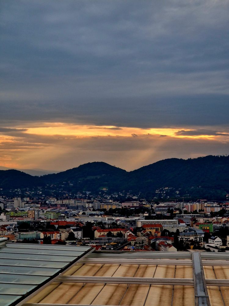 Blick auf die Dächer von Graz und zwei Hügel, hinter denen das Licht der Abendsonne die Wolken durchbricht.