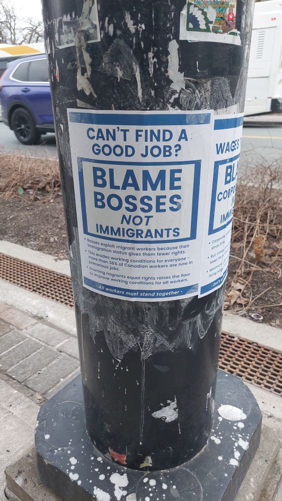 A black metal pole on a city sidewalk with a poster attached to it. The poster has a bold blue and white design with capitalized text. 

The main message reads:

"CAN'T FIND A GOOD JOB?
BLAME BOSSES NOT IMMIGRANTS"

Below this, additional text explains:

• "Bosses exploit migrant workers because their immigration status gives them fewer rights."

• "This erodes working conditions for everyone—more than 36% of Canadian workers are now in precarious jobs."

• "Granting migrants equal rights raises the floor to improve working conditions for all workers."

At the bottom, a smaller line of text states:
"All workers must stand together."

Parts of another similar poster are visible on the same pole. The background includes a street with a parked car, a bus, and some bare bushes. The pole is worn, with remnants of older stickers and peeling paint.

