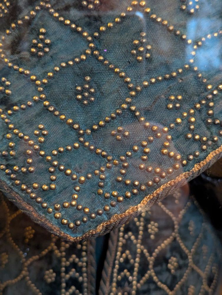 Closeup of a suit of Indian fabric and metal armor. The fabric is a dark green, and there are elaborate Islamic patterns all over the fabric, made up of tiny rivets. This is a close up of the tip of the arm piece