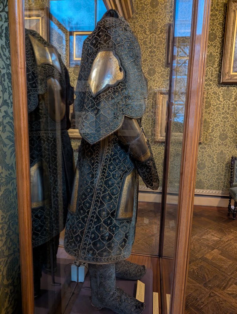 The side view of a suit of Indian fabric and metal armor. The fabric is a dark green, and there are elaborate Islamic patterns all over the fabric, made up of tiny rivets. The arm pieces cover the shoulders and hang down loose to the elbows. 