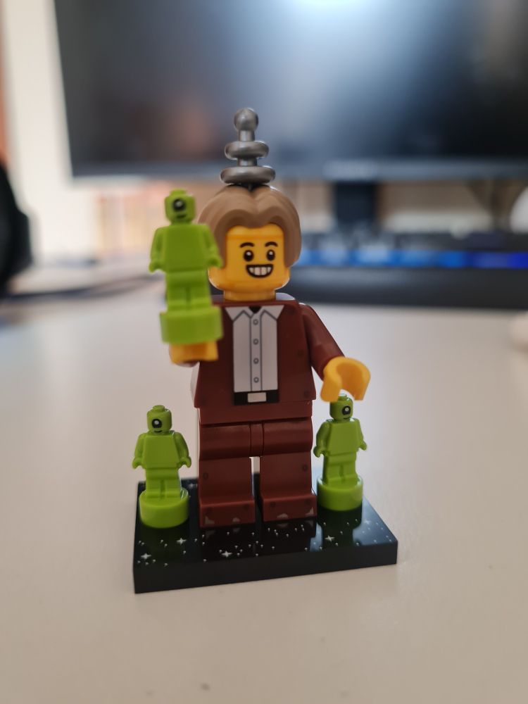 A LEGO minifigure depicting, on first glance, a totally ordinary man with dirty blonde hair parted in the middle, wearing a brown suit. However, he's standing alongside some small one-eyed green aliens (nanofigures) and holding one in his hand. An antenna sticks out the top of his head. Upon close inspection, his face is a bolted on metal plate and more suspicious bolts can be seen around the edges of his torso. A touch of metal grey can be seen on his feet, as if some brown paint has worn away revealing metal underneath.