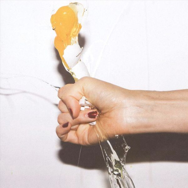 Caucasian hand with nails painted blood red squeezing a chicken's egg until it explodes out of its shell. White background, brightly lit so that there's a shadow.