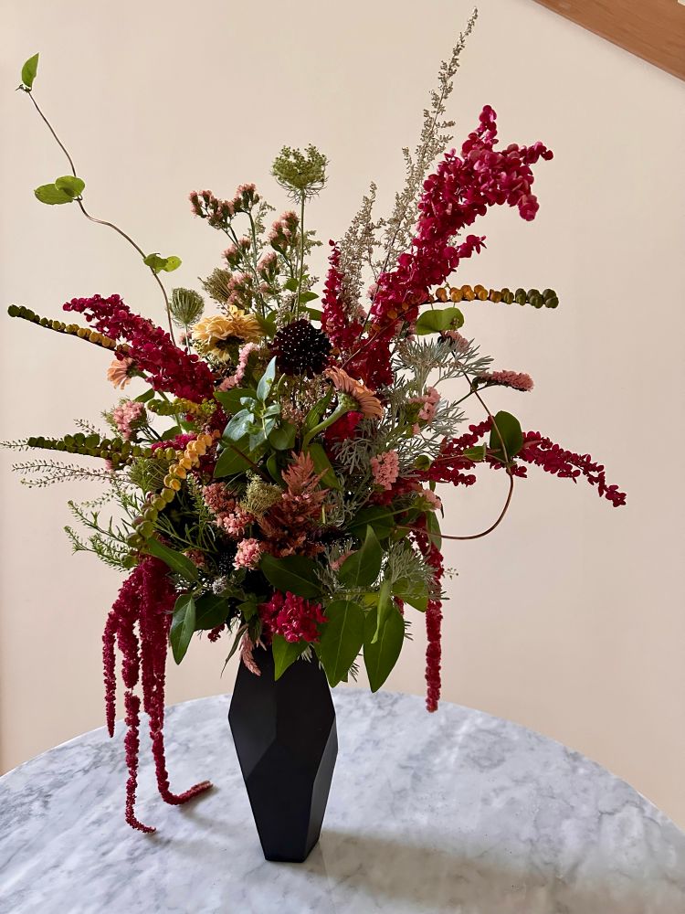 Flower arrangement with draped amaranth fronds in a black vase on a marble tabletop with a cream-colored background. 