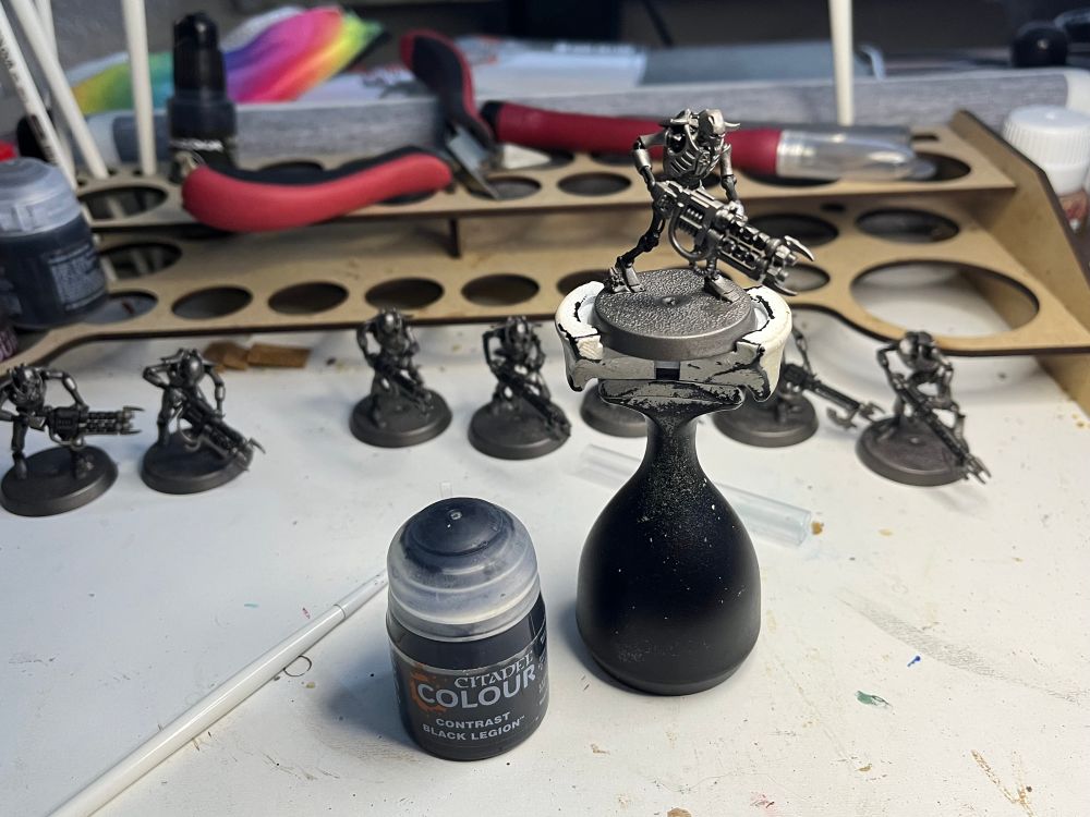 Set of Necron warrior miniatures from Warhammer 40k, one on a painting stand, next to it a pot of Color saying “contrast black legion” 