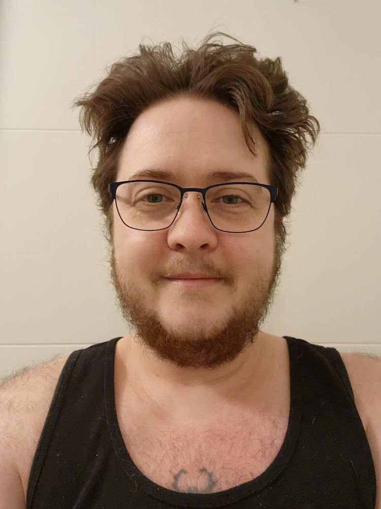 Picture of Gadfly with some new navy blue glasses, in a black tank top, with shaggy beard chest and shoulder hair. Behold his rugged masculinity!