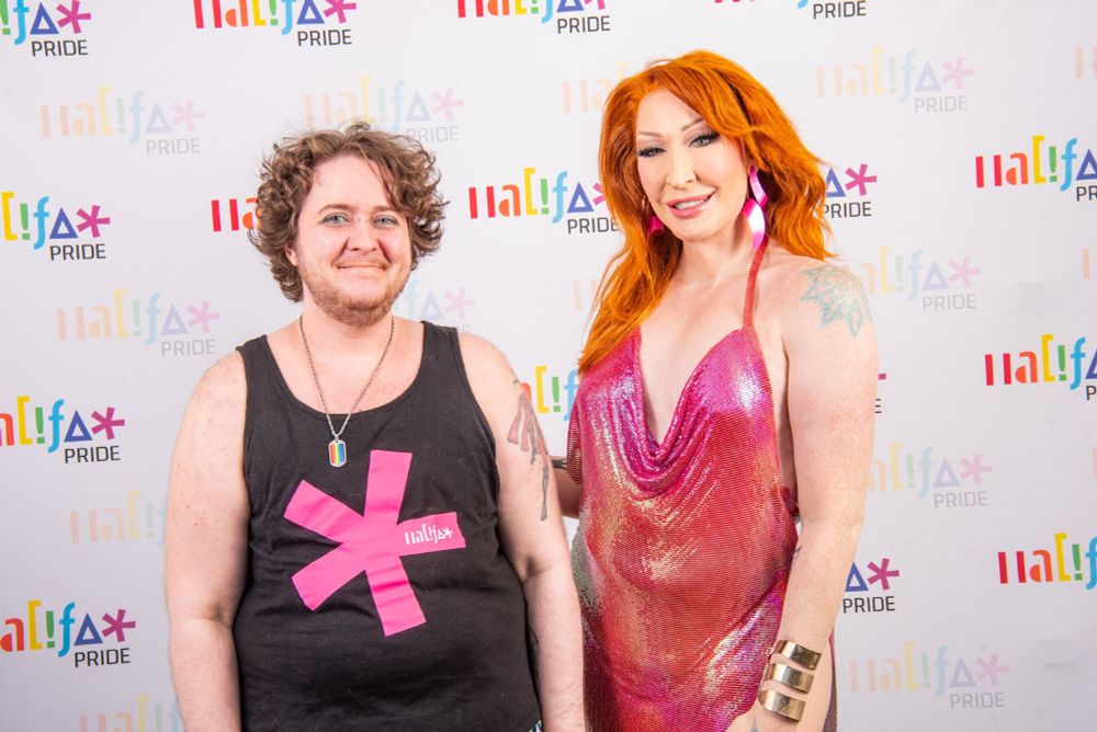 Photo of Gadfly and Detox from the Halifax Pride Meet and Greet. Detox is on the right and looks fabulous in a pink shiny dress and red hair. Gadfly is on the left and mostly looks tired and is wearing the iconic Halifax Pride butthole asterisk shirt. 