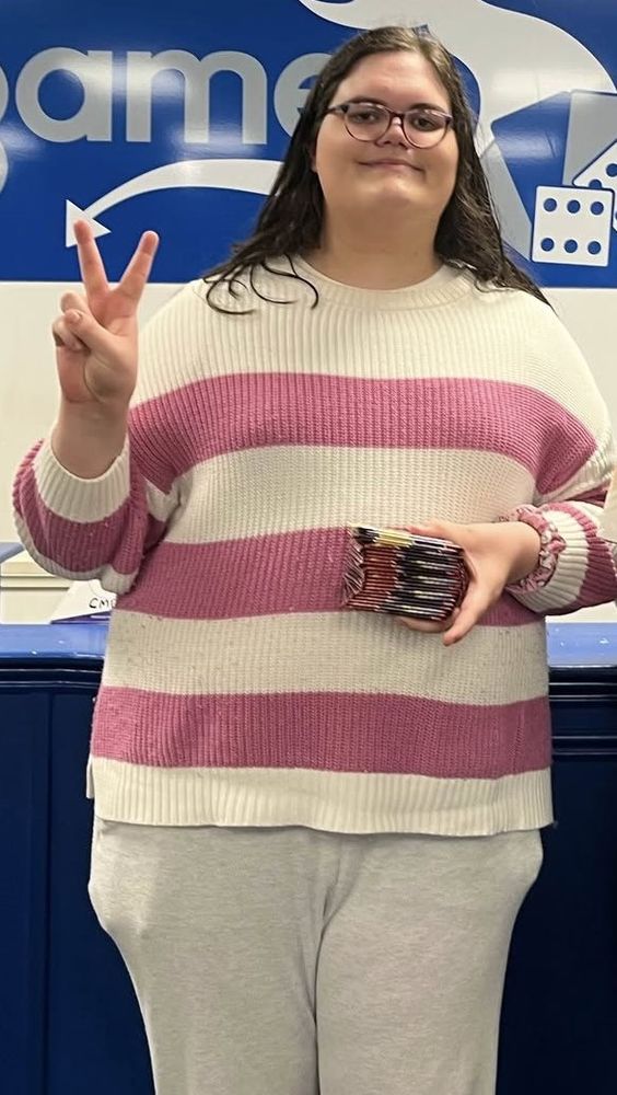 An image of me at the game store doing a peace sign with my right hand and holding the packs below my chest with the left hand.