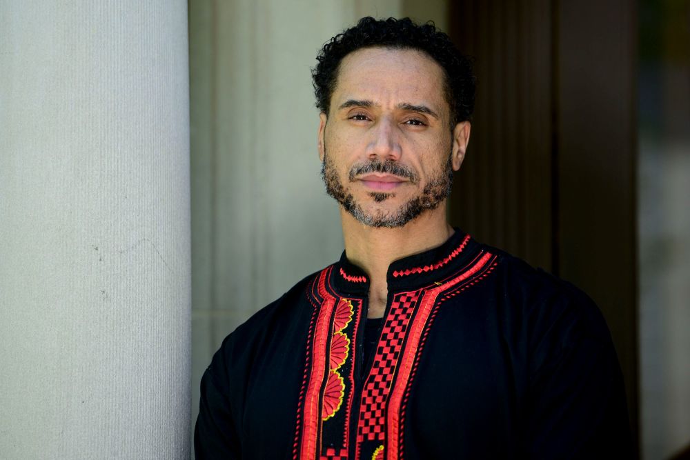 Dr. Butch Ware featured with a serious expression. Wearing a black shirt with bright red trim and patterns. Dr. Ware has dark curly hair, a mustache and some facial hair, not quite a full beard and a dark brown complexion. 