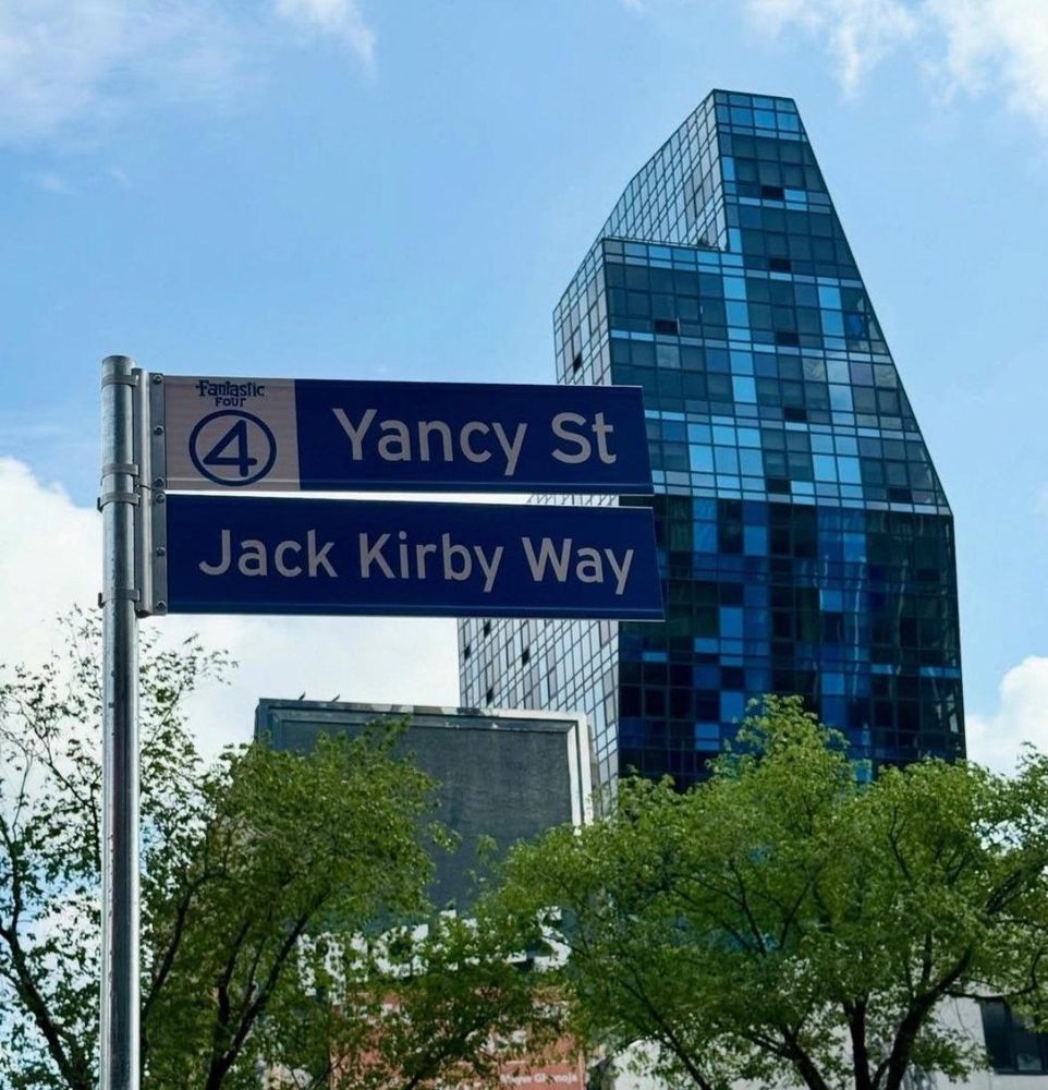 Two blue street signs in front of a high rise the signs have a fantastic four logo and say Yancy St and Jack Kirby Way