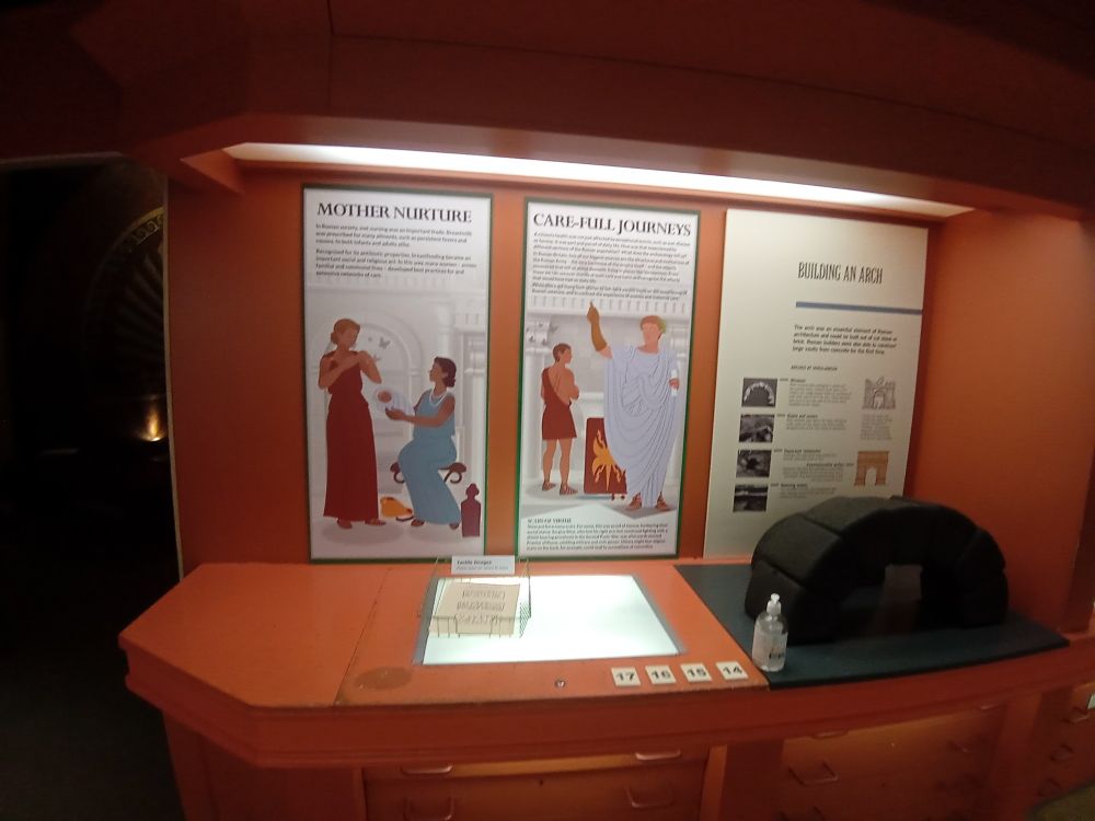 Two portrait panels on a wall with colourful illustrations. One depicts her mother handing her child to a wet-nurse. The other depicts two Roman veterans; one with a prosthetic arm, the other with scars along his back.