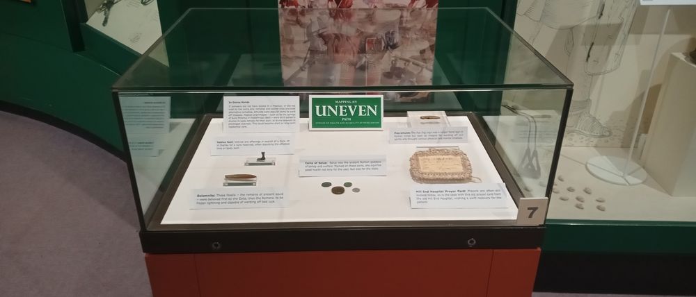 A tabletop case containing a number of museum artefacts and labels. They include a small metal votive foot, some coins depicting the Roman goddess Salus, and a 20th century prayer card from a psychiatric hospital.