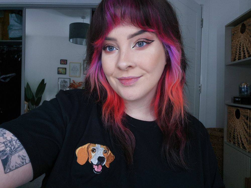 Selfie of me wearing a black t shirt, it has my late doggo stitched on the front. She's a lemon beagle and looks so happy!