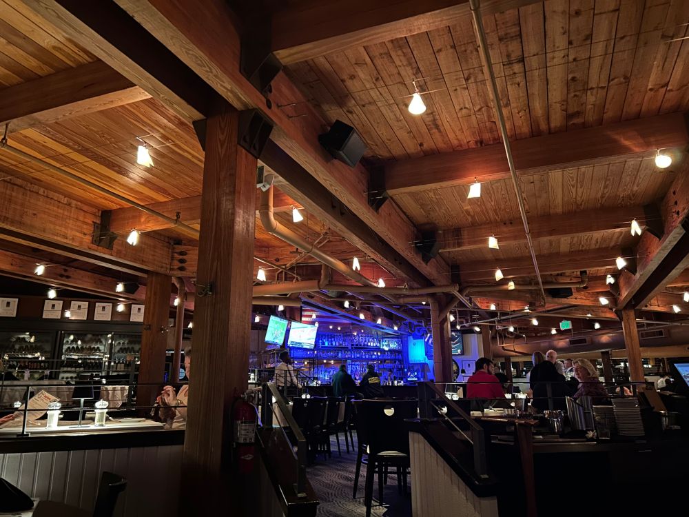 Evening view of the interior of the Rusty Scupper restaurant. Wood paneled ceiling with heavy timber beams and columns, twinkling mood lighting, and the bar is visible in the background.